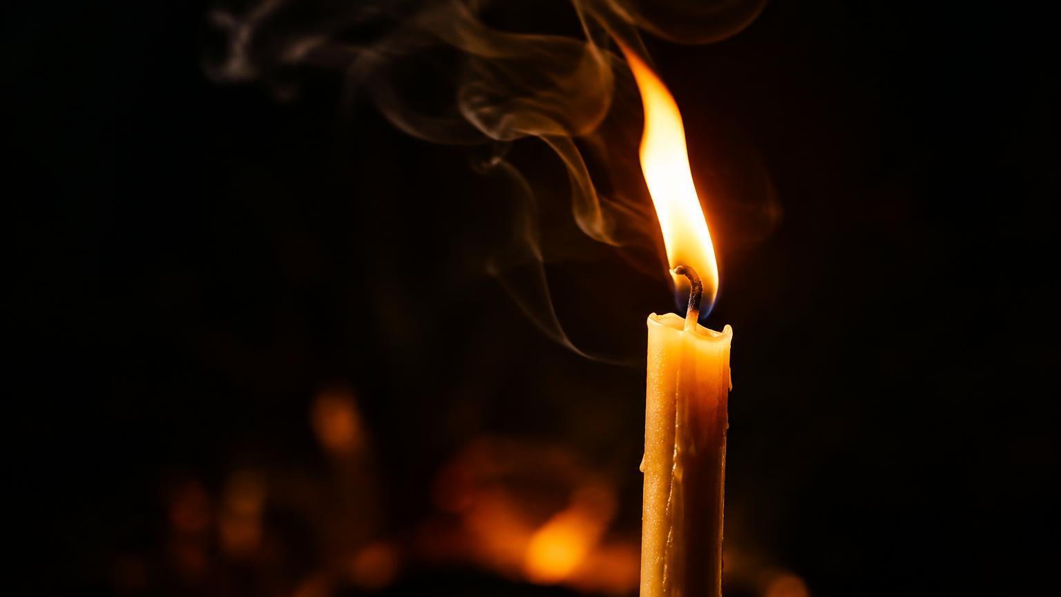 Close-up of a single beeswax taper candle burning against a dark background, its tall golden flame leaning slightly to the left as pale smoke curls upward.